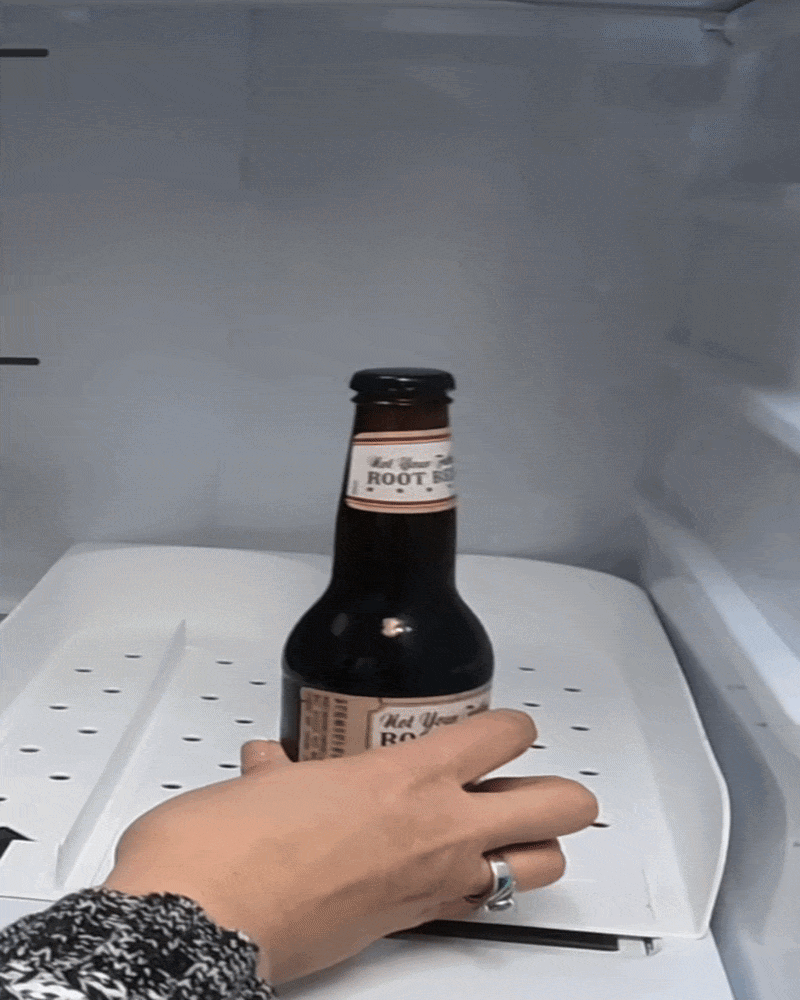 Hand placing a root beer bottle into a U-shaped fridge beverage organizer tray demonstrating FIFO drink rotation system for organized refrigerator storage