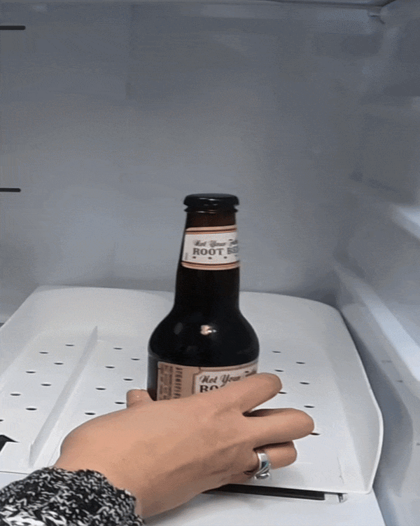Hand placing a root beer bottle into a U-shaped fridge beverage organizer tray demonstrating FIFO drink rotation system for organized refrigerator storage